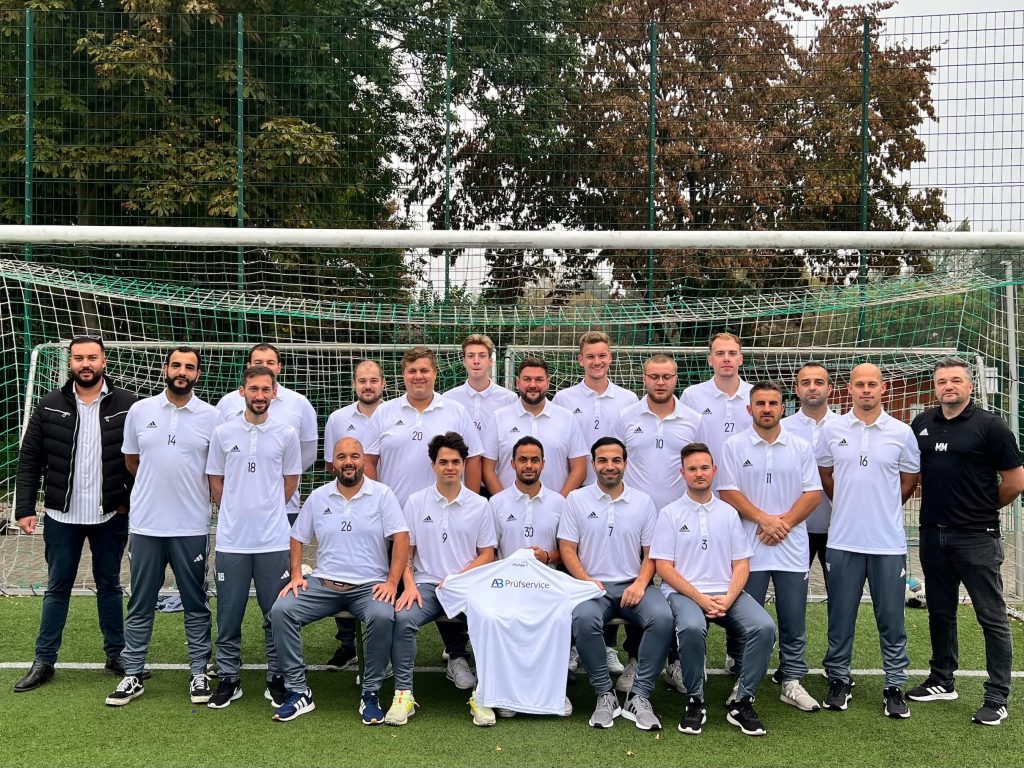 Eine lokale Fußballmannschaft posiert in passenden weißen Hemden und grauen Hosen auf einem Feld und repräsentiert den BSV Schüren. Einige knien, während andere dahinter stehen. Einer hält stolz ein Mannschaftstrikot. Im Hintergrund sind ein Tornetz und Bäume zu sehen, die die Essenz des lokalen Sports einfangen.